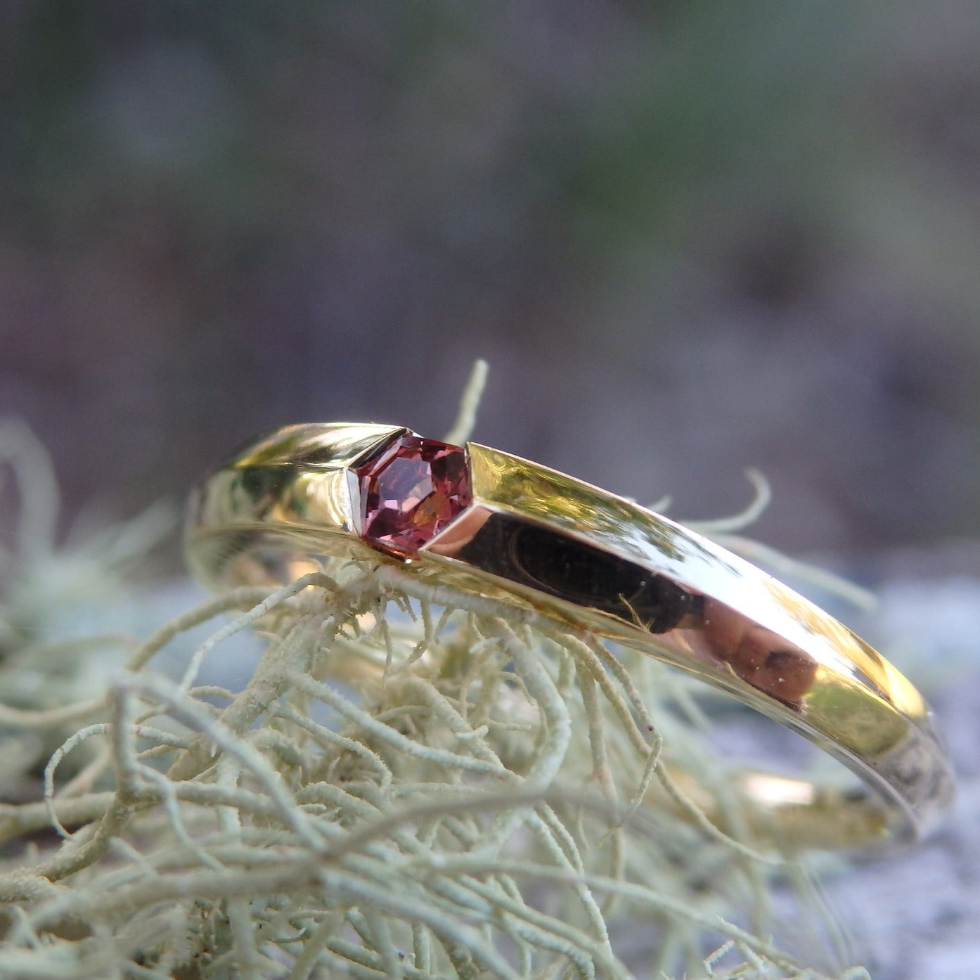 hexagon hexagonal cut spinel gemstone in a sunset-red shade sitting horizontally in a knife-edge (pointed profile) band of 18ct yellow gold, tapering from the width of the hexagon 2.5mm to 1.7mm at the back. the ring sits on a lichen plant of very pale pastel green, the reflection on the ring is my hand holding a camera