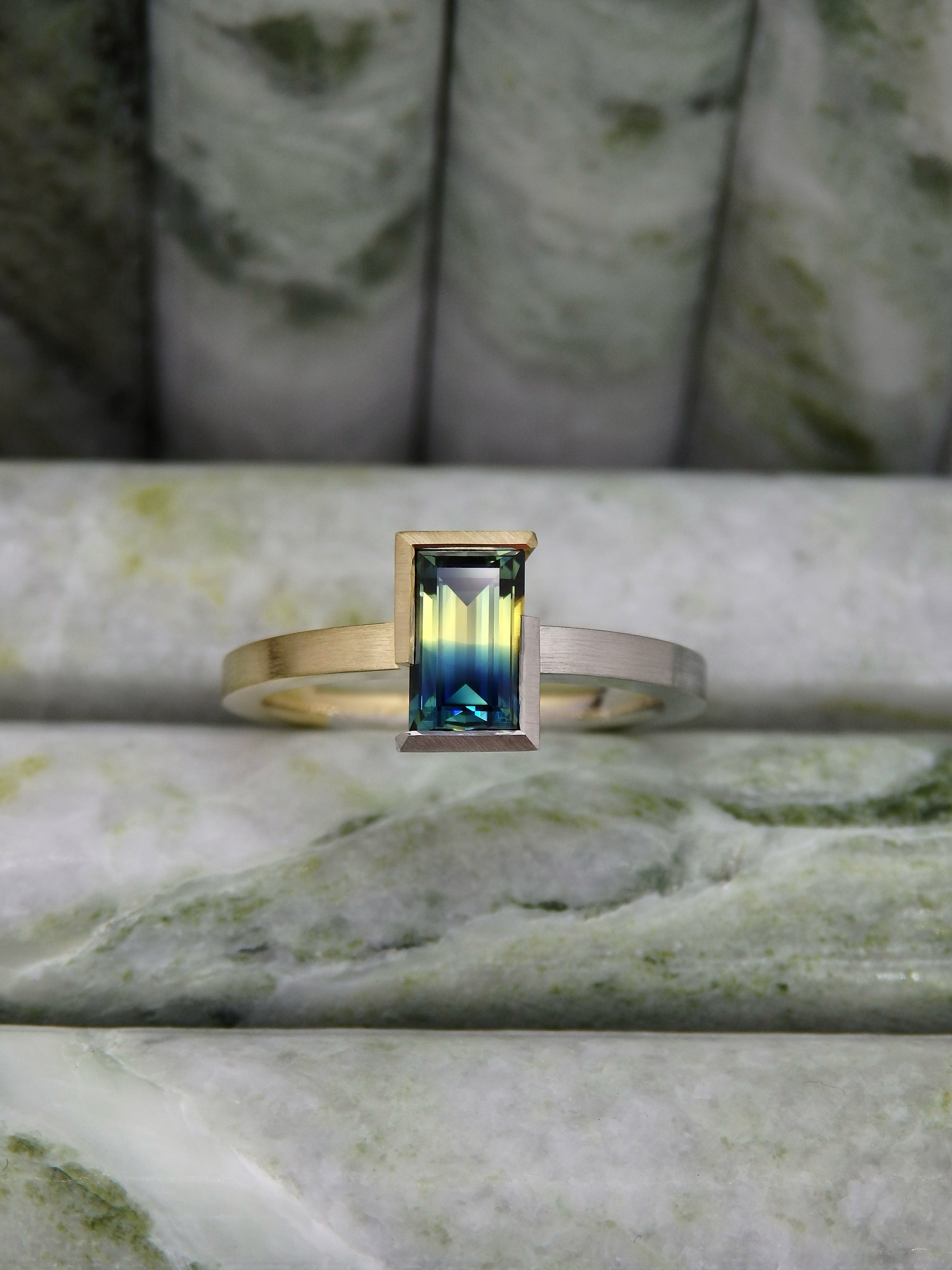 Unheated Baguette Australian Parti Sapphire, a perfectly split crystal of yellow and blue held in a geometric two-tone wrap around semi-bezel, one half of the ring made from platinum and the other half 18ct yellow gold. 
Displayed on green marble background 