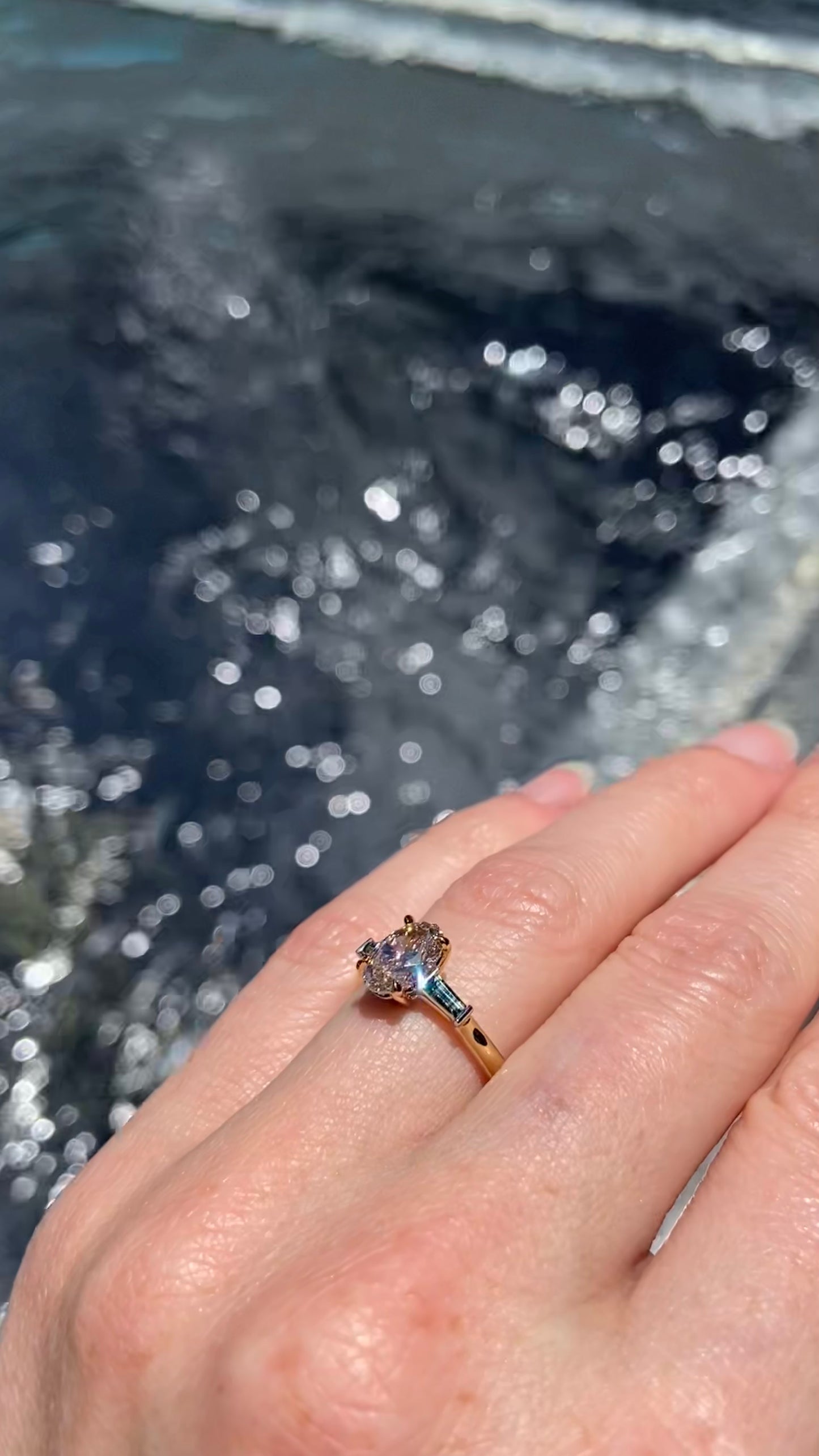 Hand wearing an engagement ring with a natural brown diamond with australian parti sapphires against a blurred water background, sparkling in the sun