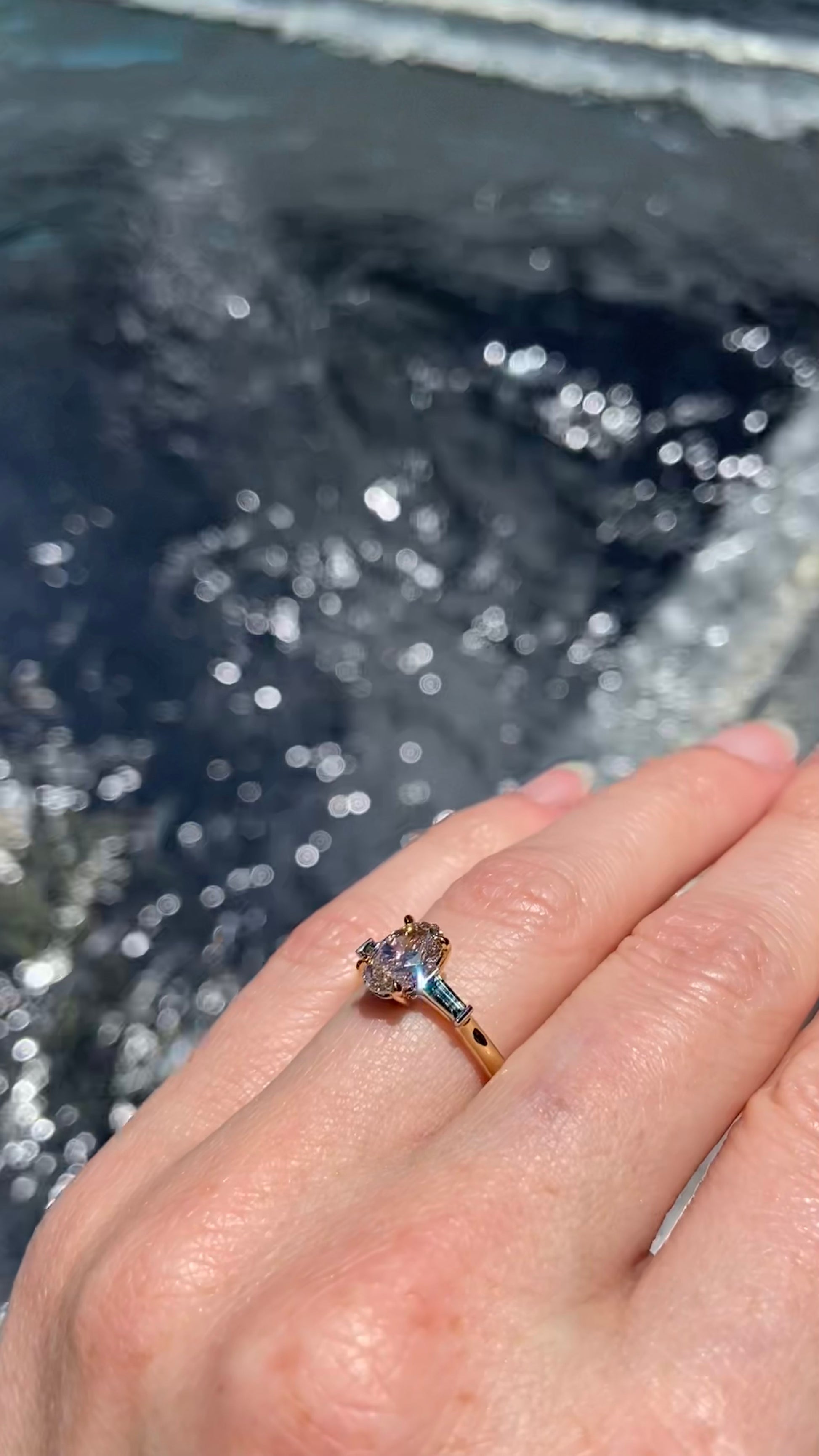 Hand wearing an engagement ring with a natural brown diamond with australian parti sapphires against a blurred water background, sparkling in the sun