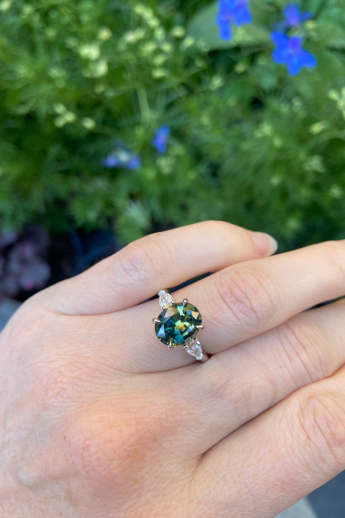 Shown on the hand in natural daylight Big oval teal parti sapphire trilogy engagement ring with natural pear shape diamond side stones handmade in platinum