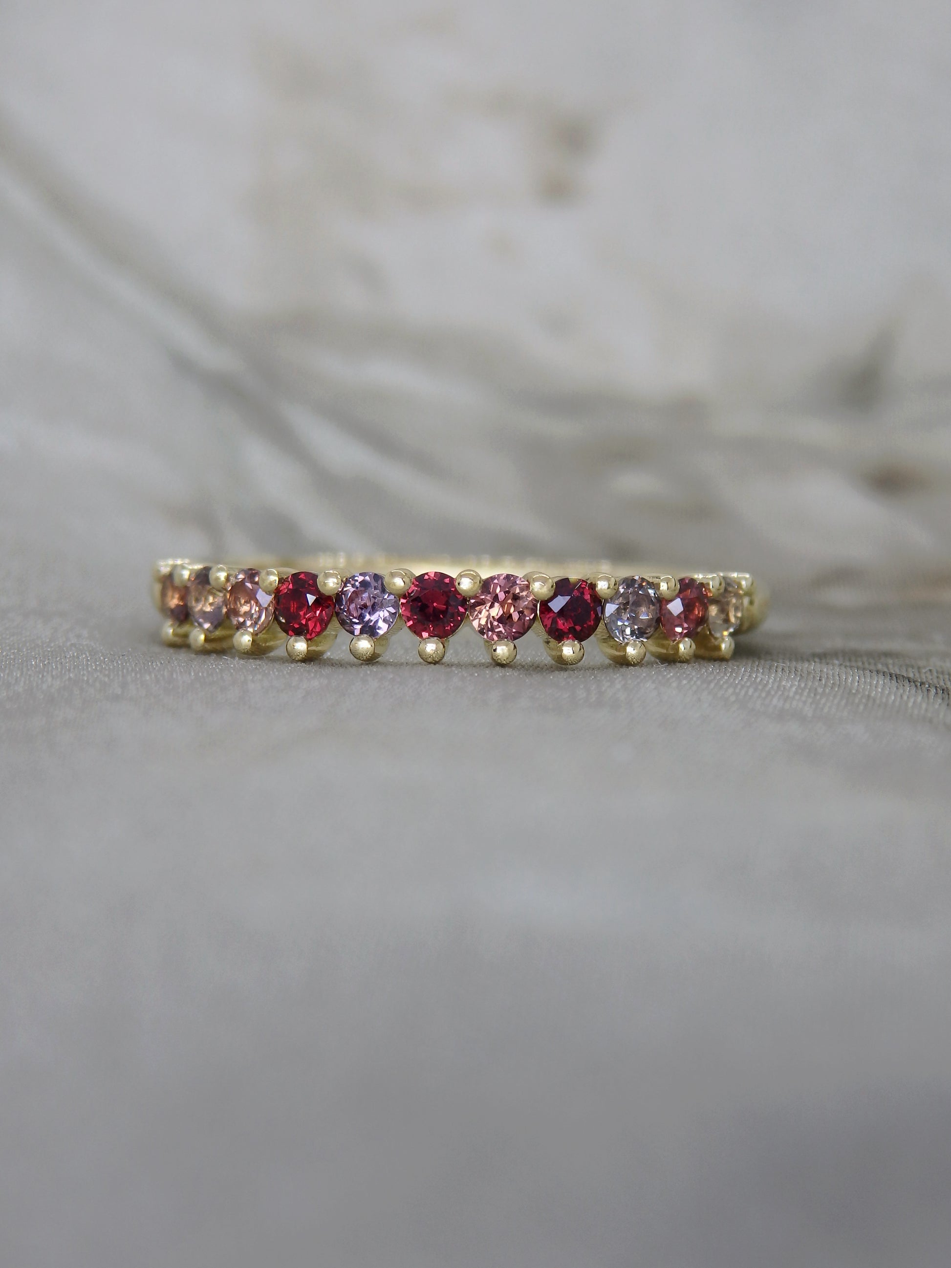 A delicate yellow gold eternity ring featuring a row of alternating pink and purple round-cut gemstones, resting on a soft gray fabric surface.