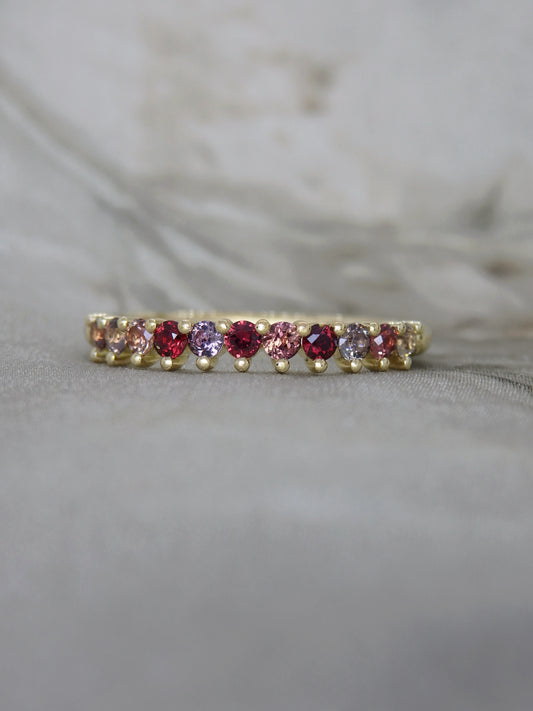 A delicate yellow gold eternity ring featuring a row of alternating pink and purple round-cut gemstones, resting on a soft gray fabric surface.