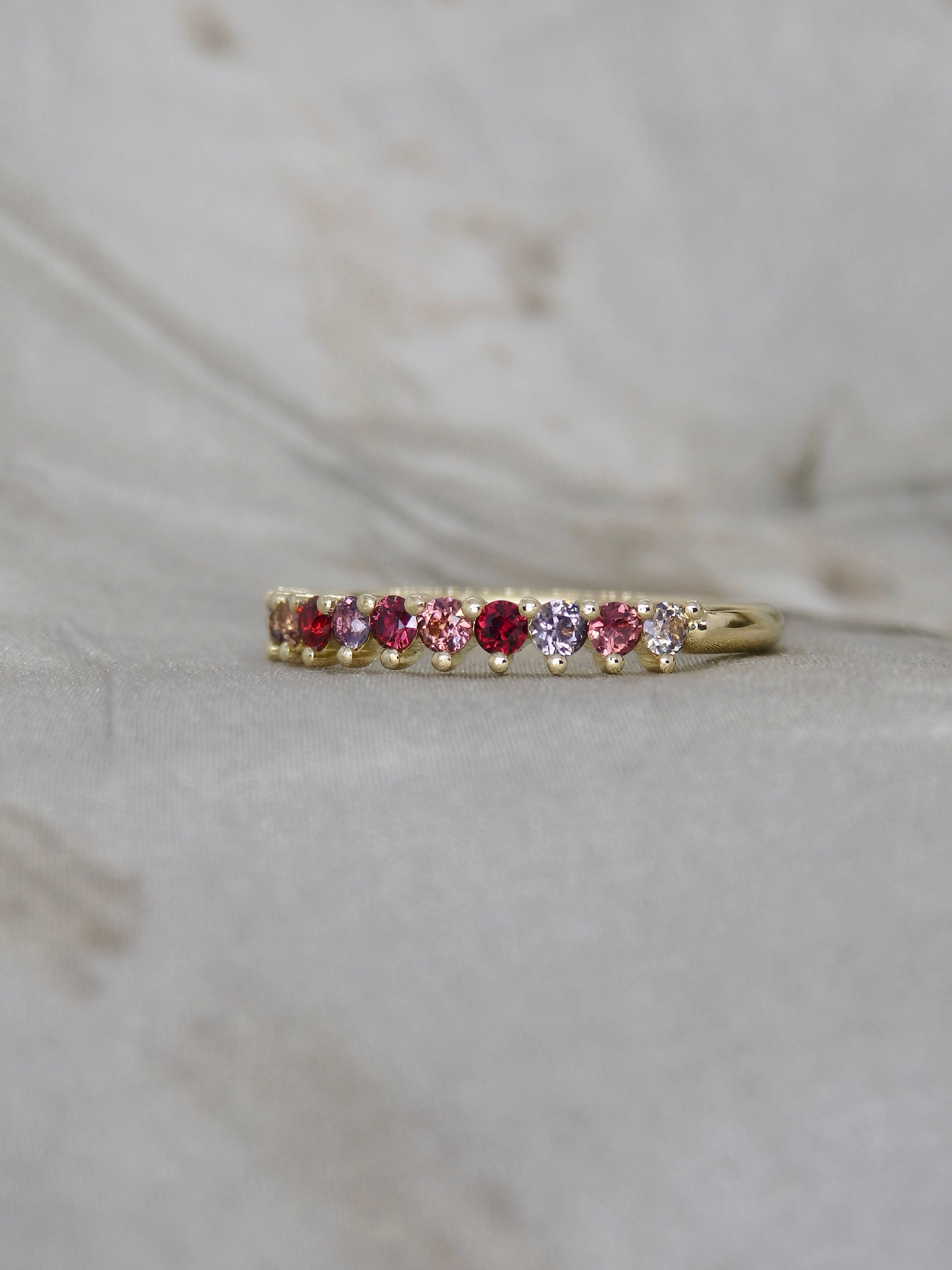 A gold eternity ring featuring a row of round-cut gemstones in red, pink, and clear, resting on a light gray marble surface.